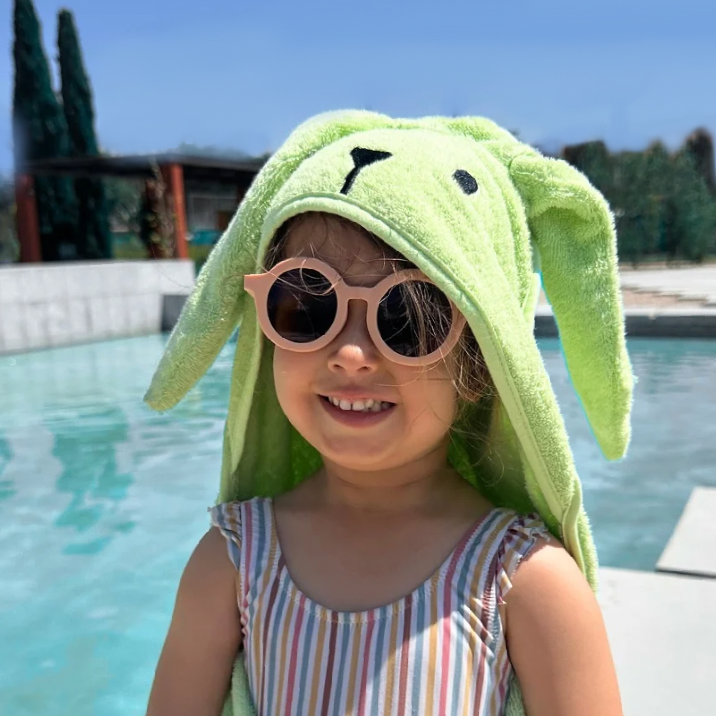 Child with a green towel wrapped around them, wearing sunglasses by a pool.
